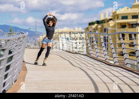 Afro Frisur Frau auf Rollschuhe Reiten im Freien auf städtischen B.. Junge fit schwarze Frau auf Rollschuhe Reiten im Freien auf Stadtbrücke mit offenen Armen. Lächelndes Mädchen mit afro Frisur Rollerblading an sonnigen Tag. Schöne Wolken am Himmel. Stockfoto