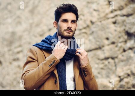 Junger Mann mit Winterkleidung in der Straße. Junge Bartgeier Kerl mit modernen Frisur mit Mantel, Schal, blaue Jeans und T-Shirt. Stockfoto