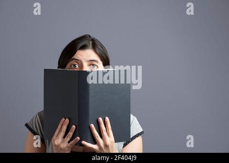 Junge Frau, die die Kamera anschaut und hinter einem offenen schwarzen Buch, das in beiden Händen gehalten wird, herausschaut. Hochformat mit grauem Hintergrund und Kopierbereich. Konzept von Stockfoto