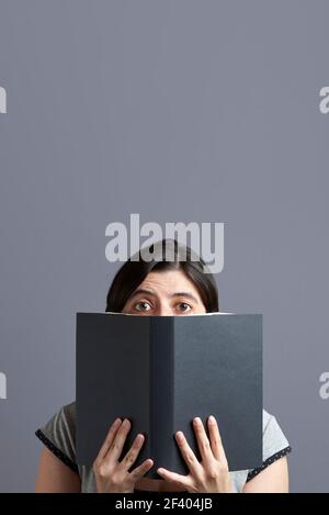 Junge Frau, die die Kamera anschaut und hinter einem offenen schwarzen Buch, das in beiden Händen gehalten wird, herausschaut. Hochformat mit grauem Hintergrund und Kopierbereich. Konzept von Stockfoto