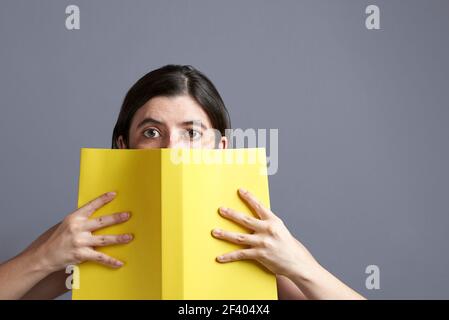 Junge Frau, die die Kamera anschaut und hinter einem offenen gelben Buch, das in beiden Händen gehalten wird, herausschaut. Hochformat mit grauem Hintergrund und Kopierbereich. Konzept o Stockfoto