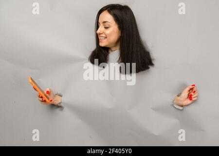 Konzept Foto von jungen weiblichen Zerreißen Papier und guckt aus Loch, hält Smartphone, isoliert grauen Hintergrund Stockfoto