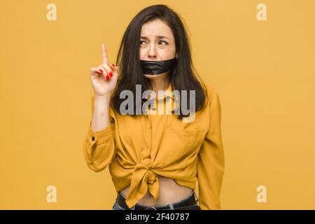 Konzeptfoto des weiblichen Opfers mit aufgeklebtem Mund. Trägt gelbes Hemd, isolierte gelbe Farbe Hintergrund Stockfoto