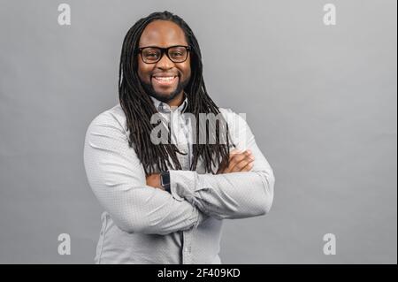 Selbstbewusst fröhlich afroamerikanisch mit langen Dreadlocks tragen smart casual Trikot steht mit gekreuzten Armen und schaut auf die Kamera mit toothy Lächeln, vielfältige Büroangestellte Konzept, isoliert Stockfoto