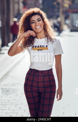 Lächelnden jungen arabischen Frau mit schwarzen Locken Frisur. Arabische Mädchen in Freizeitkleidung in der Straße. Gerne weibliche tragen weiße T-Shirt und Hose. Stockfoto