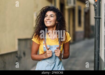 Junge arabische Touristen Frau mit schwarzen Locken Frisur im Freien. North African Reisenden Mädchen in lässiger Kleidung auf der Straße. Glückliche Frau trägt gelbe T-Shirt und Denim-Kleid im städtischen Hintergrund. Stockfoto