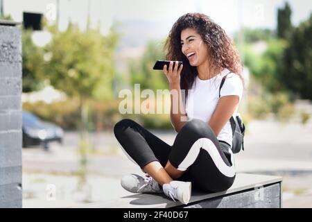 Schöne afrikanische Frau Aufzeichnung einer Sprachnotiz mit Smart Phone im Freien. Arabische Mädchen im Sport Kleidung mit Curly Frisur im städtischen Hintergrund. Stockfoto