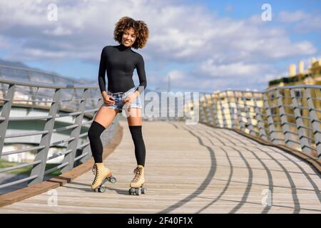 Schwarze Frau, afro Frisur, auf Rollschuhen reiten im Freien auf städtischen Brücke mit offenen Armen. Lächelnde junge weibliche rollerblading an einem sonnigen Tag. Schöne Wolken am Himmel. Stockfoto