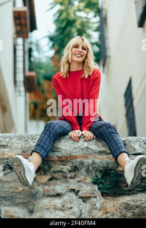 Gerne blonde junge Frau sitzt auf der städtischen Hintergrund. Lächelnde blonde Mädchen mit roten Hemd, genießen das Leben im Freien. Stockfoto