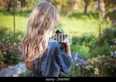 Kleines Mädchen mit langen blonden Haaren hält schwarze Katze Baby auf der Schulter, Rückansicht in den bunten Garten während des sonnigen Tages Stockfoto