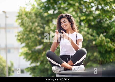 Schöne afrikanische Frau, die Musik mit Ohrhörern und Smartphone im Freien hört. Arab Mädchen in Sportkleidung mit lockigen Frisur im städtischen Hintergrund. Stockfoto