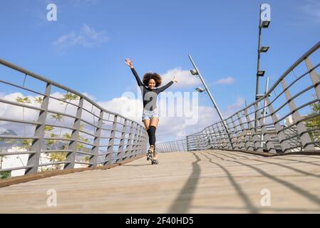 Schwarze Frau, Afro Frisur, auf Rollschuhe Reiten im Freien auf Stadtbrücke mit offenen Armen. Lächelnd junge weibliche Rollerblading an sonnigen Tag. Schöne Wolken am Himmel.. Afro Frisur Frau auf Rollschuhe Reiten im Freien auf der städtischen Brücke Stockfoto