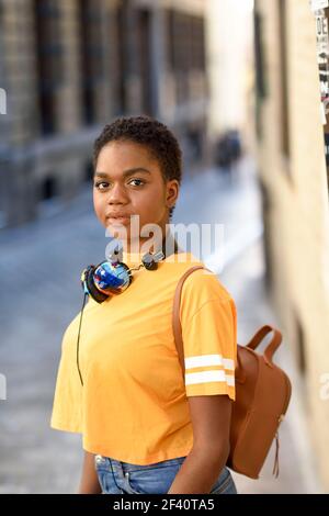 Junge afrikanische Frau in legerer Kleidung und Kopfhörer Blick auf die Kamera im städtischen Hintergrund. Lifestyle-Konzept... Junge afrikanische Frau in legerer Kleidung und Kopfhörer Blick auf die Kamera Stockfoto