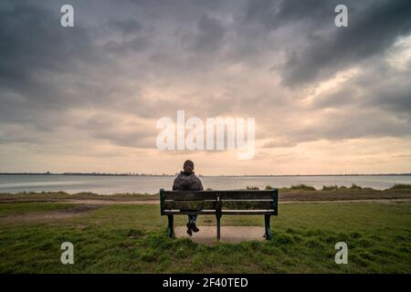 Rückansicht einer Frau, die auf einer Bank sitzt und auf das Meer blickt, launischer Himmel. Stockfoto