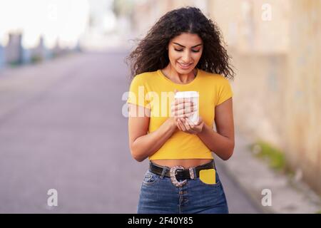 Junge arabische Frau, die mit einem Kaffee zum Mitnehmen über eine urbane Straße geht. Arabisches Mädchen, das mit einem Kaffee zum Mitnehmen über die Straße geht Stockfoto