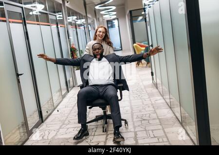 Überglücklich afroamerikanischer Mann mit kaukasischen Frau, die ein Rennen auf Stuhl in der Bürohalle macht, Spaß zu haben. Freundliche, multirassische Kollegen machen eine Pause vom Arbeits-, Freizeit-Konzept Stockfoto