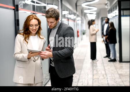 Elegante Geschäftsleute stehen im Büro und nutzen ein Tablet. Hübscher männlicher Angestellter, der einen Rat von der schönen Rothaarige Frau Kollegin über Arbeit Fragen fragt Stockfoto