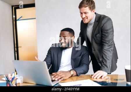 Freundliche, multiethnische männliche Kollegen, die gemeinsam an einem Unternehmensprojekt arbeiten, mit einem Laptop. Erfolgreiche Geschäftsleute in stilvollen formellen Anzügen auf der Suche nach Ideen für Startup, Lächeln, Partnerschaftskonzept Stockfoto