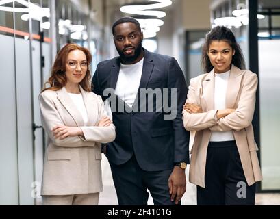 Einflussreiche Geschäftspartner blicken selbstbewusst und lächelnd auf die Kamera. Multirassische erfolgreiche Kollegen in formaler stilvoller Kleidung stehen im modernen Büro, Teamwork-Konzept Stockfoto