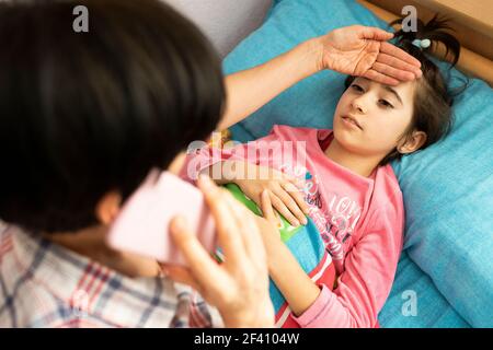 Mutter besorgt über ihre Tochter&rsquo;s Temperatur ruft den Arzt. Kleines Mädchen auf dem Bett. Mutter besorgt über ihre Tochter&rsquo;s Temperatur ruft den Arzt Stockfoto
