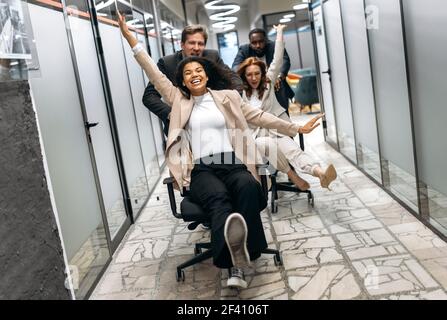 Glückliche freundliche Mitarbeiter in formeller Kleidung machen Rennen auf Stühlen auf Freizeit im Büro. Multiethnische Geschäftspartner haben Spaß während der Arbeitspause, lächeln und lachen Stockfoto
