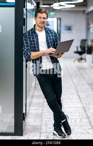 Kaukasion männliche Freiberufler steht in modernen Büro, Blick auf die Kamera, lächelnd. Happy junge Erwachsene Geschäftsmann in stilvoller Kleidung ist mit Laptop, arbeiten an neuen Projekt Stockfoto