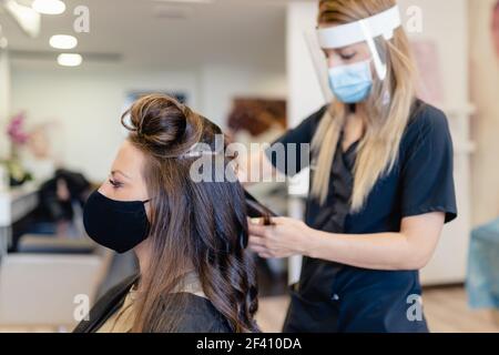 Friseur, durch eine Maske geschützt, macht Wellen in ihrem Client&rsquo;s Haar mit einem Haareisen in einem Salon. Business- und Beauty-Konzepte. Friseur, durch eine Maske geschützt, Kämmen ihrer Client&rsquo;s Haar mit einem Haareisen in einem Salon. Stockfoto
