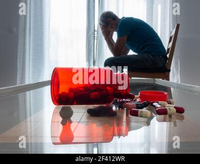 Reifer Mann (63 Jahre alt) mit Kopf in den Händen in der Nähe des Fensters mit einer Flasche Pillen auf dem Tisch. Männliche Depression, psychische Gesundheit, psychische Krankheit... Konzept Stockfoto