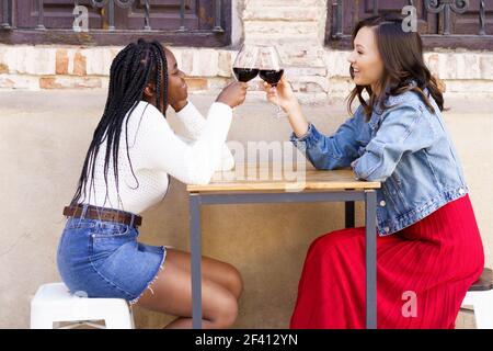 Zwei Freunde machen einen Toast mit Rotwein an einem Tisch vor einer Bar sitzen. Multiethnische Frauen... Zwei Frauen machen einen Toast mit Rotwein an einem Tisch vor einer Bar sitzen. Stockfoto