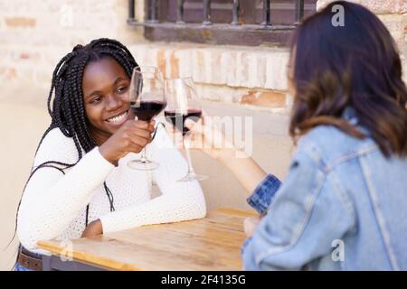 Zwei Freunde machen einen Toast mit Rotwein an einem Tisch vor einer Bar sitzen. Multiethnische Frauen... Zwei Frauen machen einen Toast mit Rotwein an einem Tisch vor einer Bar sitzen. Stockfoto