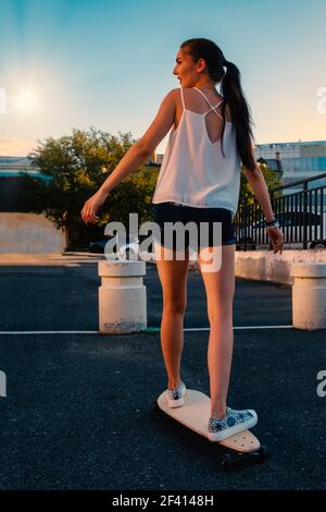 Schönes Mädchen Reiten Skateboard in kurzen Denim-Shorts, Blick von hinten vor dem frühen Abendhimmel. Schöne Mädchen Reiten Skateboard in kurzen Denim-Shorts, Blick von hinten Stockfoto