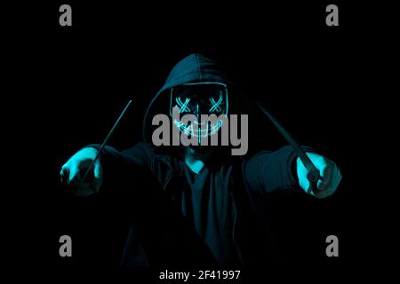 Mann bewaffnet mit zwei Macheten und trägt eine gruselige Beleuchtung Neon Glow Maske und ein Hoodie auf schwarzem Hintergrund. Stockfoto