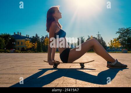 Schöne junge Mädchen in kurzen Shorts und Sport Tank Top sitzt auf Skateboard bei sonnigem Wetter durch Tageslicht mit Zickzack Schatten auf Bürgersteig hinterleuchtet. Schöne junge Mädchen in kurzen Shorts und Sport Tank Top sitzt auf Skateboard bei sonnigem Wetter Stockfoto