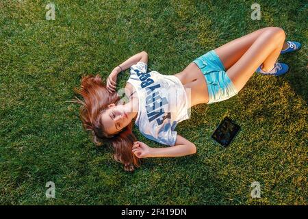 Junge Frau auf Gras mit Tablet-Computer in der Nähe liegen und lange Ingwer Haare über den Kopf verteilt. Junge Frau, die auf Gras ruht, mit Tablet-Computer und langen Ingwerhaaren, die über den Kopf verteilt sind Stockfoto