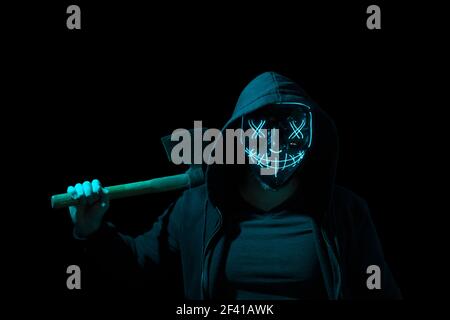 Mann bewaffnet mit einer Axt und trägt eine gruselige Beleuchtung Neon Glow Maske und ein Hoodie auf schwarzem Hintergrund. Stockfoto