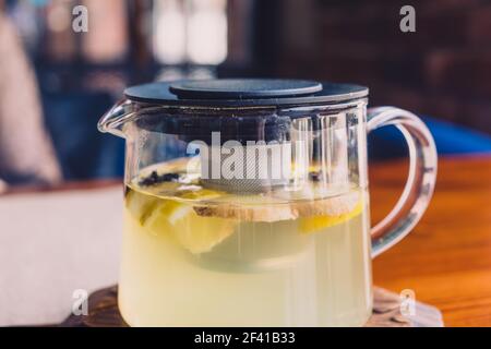 Heißer Kräutertee befindet sich in einem Glaskessel auf der Tischplatte in der Cafeteria. Auf der Oberfläche des Getränks schweben Stücke von Ingwer, Zitrone und Zimt. Kräutertee ist in einem Glaskessel. Stockfoto
