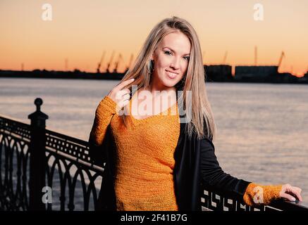 Glückliche Frau in orange gestrickter Jacke posiert auf Böschung vor der Skyline der Dämmerung auf Zaun gelehnt. Fröhliche Frau in orangefarbener Strickjacke, die vor der Skyline von Dawn auf dem Damm posiert Stockfoto