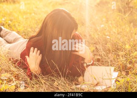 Mädchen liest im herbstlichen Park, Bild hinterleuchtet, sie schaut auf Buch und ihr Gesicht ist durch ihre Haare versteckt Stockfoto