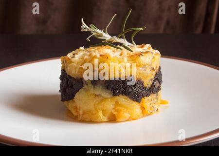 Nahaufnahme von gerösteten Kartoffeln, Blutwurst und Käsekuchen, garniert mit einem Zweig frischen Rosmarins auf einem braunen und weißen Keramikplatte. Stockfoto