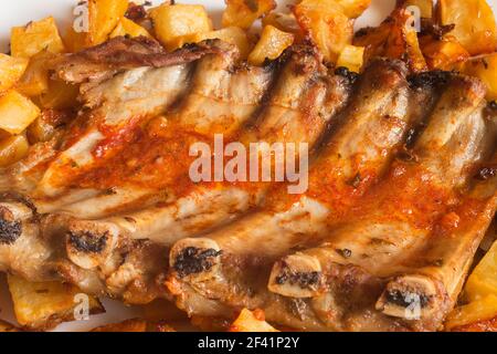 Detail von gebackenen Schweinefleischrippen mit Knoblauchsauce, Öl, Essig und würzigen Gewürzen umgeben von Kartoffeln. Frische Lebensmittel und Grillfleisch. Stockfoto