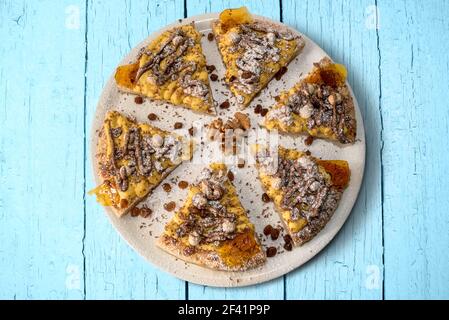 Süße Pizza mit Schokolade Haselnusscreme, Eiercreme, Rosinen und Nüssen, sechs Scheiben in weißem Teller auf hellblauen Holzbrettern, flach in Draufsicht Stockfoto