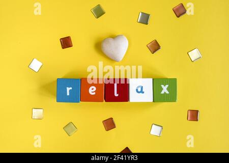 RELAX - Wort aus farbigen Holzblöcken. Bemalte Würfel mit Buchstaben und einem Steinherz auf gelbem Hintergrund. Draufsicht. Speicherplatz kopieren. Stockfoto