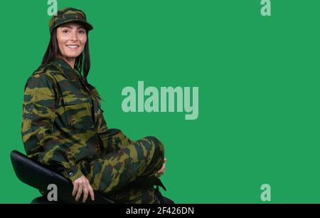 Porträt einer schönen kaukasischen Brünette Frau in Militärhut und Uniform, lächelnd, posiert niedlich auf einem Stuhl sitzen. Seitliche Leerstelle für Werbung Stockfoto