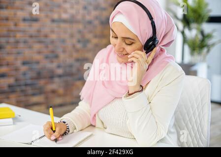 Lächelnde arabische junge Frau verwendet Headset, Blick auf Notebook, Hören Lernen Online-Kurse. Glückliche religiöse Geschäftsfrau mit Kopfhörern, Videoanruf für Kundenservice berühren den Hörer Stockfoto