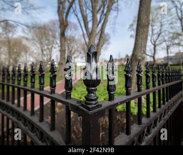 Ein schmiedeeiserner Zaun an einem Park. Blick ist auf eine Ecke des Zauns. Bäume und ein Ziegelweg sind im Hintergrund. Stockfoto