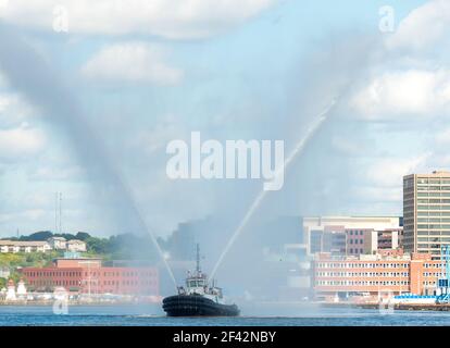 Schleppboot im Hafen feuern. Schlepper sprüht zwei Wasserströme in V-Form in die Luft. Schlepper teilweise durch Spray verdeckt. Stadt im Hintergrund. Stockfoto