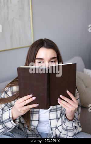 Fröhliche Frau bedeckt Gesicht mit altem Buch, hat freudigen Ausdruck, in hohem Geist zu sein, wie ihre Lieblingsgeschichte liest, mag Fiktion, verbringt Freizeit mit Stockfoto