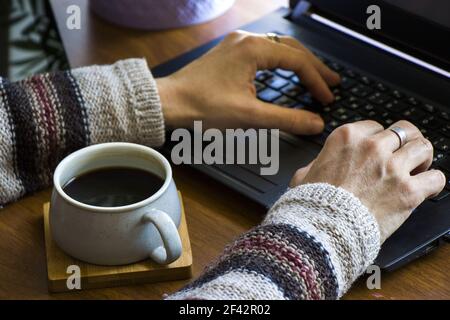 Eine Tasse Kaffee und weibliche Hände, die auf dem Laptop tippen; Arbeitsprozess und Arbeitsplatz zu Hause während der Quarantäne Stockfoto