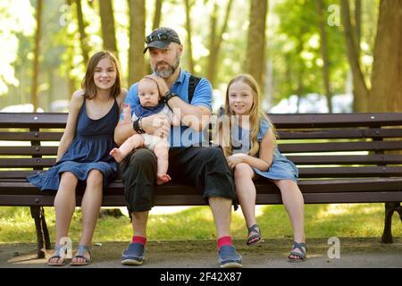 Vater und drei Kinder Spaß am Sommertag im Stadtpark. Liebenswert Baby Junge wird von seinem Papa gehalten. Zwei ältere Schwestern umarmen ihren Vater und BA Stockfoto