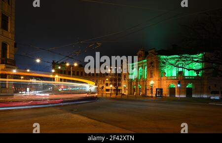 Die Straßen der Stadt sind nachts durch kurvenförmige Lichtwege zu erreichen, während die Straßenbahn vorbeifährt. Einzigartiges grün beleuchtetes Gebäude im Hintergrund. Stockfoto